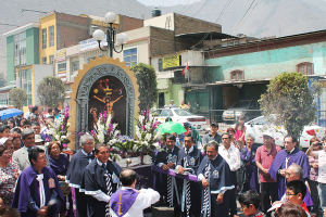 Menschen auf einer Stra├če mit einem Kruzifix und Blumenstr├Ąu├čen in der Mitte, Autos im Hintergrund und Geb├Ąude, B├Ąume und Stra├čenlaternen dahinter.