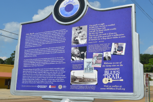 Ein Informationsschild, Gebude, ein Schuppen, Bume, Stromkabel und ein bewlkter Himmel.