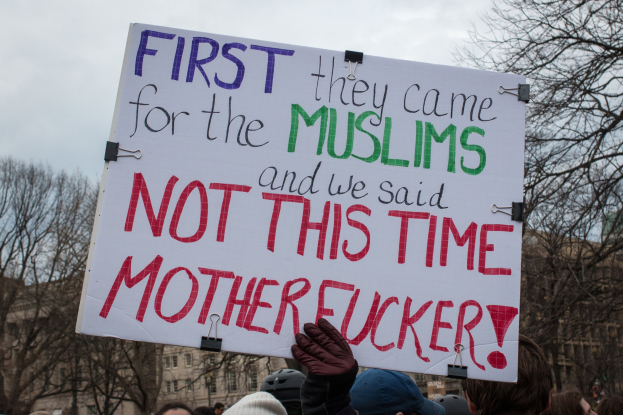 Eine Gruppe von Menschen mit Möppen und Handschuhen hölt ein Schild mit der Aufschrift "Zuerst kamen sie wegen der Muslime und wir sagten nicht dieses Mal, verdammte Scheiße" mit Bäumen, Gebäuden und einem klaren blauen Himmel im Hintergrund.
