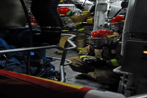 Feuerwehrleute in Schutzausrüstung arbeiten an einem Löschfahrzeug mit Rohren und anderen Gegenständen im Hintergrund.