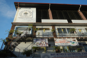 Mariä-Mutter-Gutenberg-Schule mit Fenstern, Geländern, Treppen, Blumentöpfen, Bannern und einem bewölkten Himmel im Hintergrund.