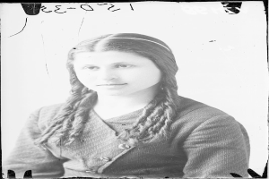 Ein Schwarz-Weiß-Porträt von Julia Margaret Cameron, eine Frau mit langen Zöpfen, die zu einem Knoten gebunden sind und ein weißes Kleid sowie einen gemusterten Schal trägt, mit einem sanften Gesichtsausdruck und ihren Händen vor der Brust verschränkt.