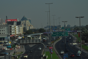 Eine Luftaufnahme einer Stadt mit Gebäuden, Türmen, Kraftfahrzeugen, Werbetafeln, Schildern, Straßen, Bäumen, Straßenlaternen, Menschen, Verkehrskegeln und Informationsschildern unter einem klaren Himmel.