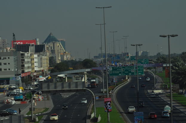 Eine Luftaufnahme einer Stadt mit Gebäuden, Türmen, Kraftfahrzeugen, Werbetafeln, Schildern, Straßen, Bäumen, Straßenlaternen, Menschen, Verkehrskegeln und Informationsschildern unter einem klaren Himmel.