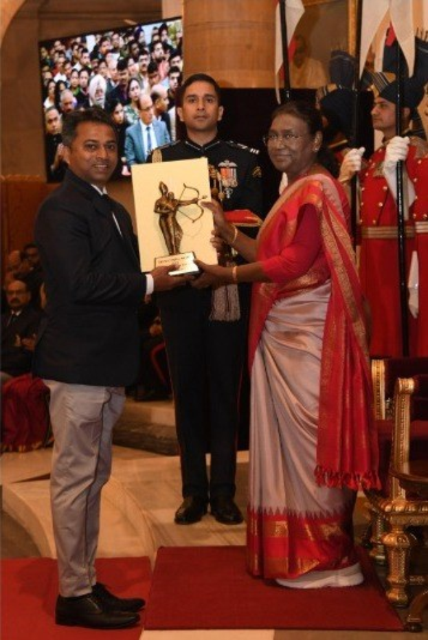 Ein Mann in einem schwarzen Blazer, weißem Hemd und grauen Hosen, der eine Trophäe hält, neben einer Frau in einem ashesfarbenen Sari auf einem roten Teppich bei einer Preisverleihung, mit Menschen, Stühlen, Fahnen und einem Bildschirm im Hintergrund.