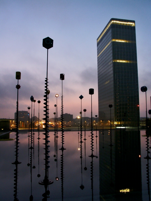 Mehrere Pfähle und Gebäude mit Lichtern, unter einem bewölkten Himmel, mit Spiegelungen der Pfähle