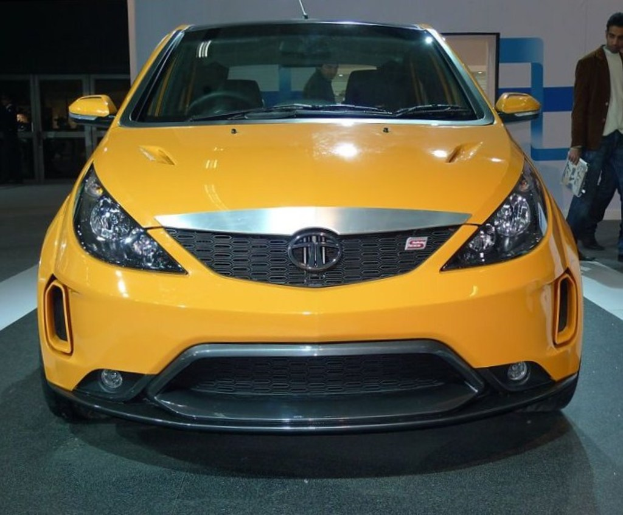 Ein oranges Auto in einer Ausstellungshalle mit einem Mann daneben, der Papiere hält, und einer Wand im Hintergrund.