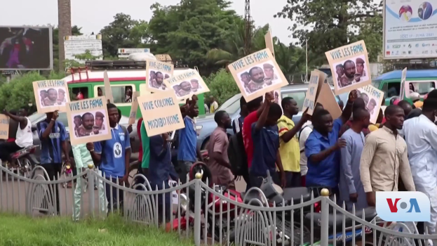 Eine Gruppe von Menschen hält Schilder und versammelt sich vor einem Zaun, umgeben von Fahrzeugen, Bäumen, Pfählen und Plakaten, mit einem Bildschirm und Gras im Hintergrund, wahrscheinlich protestieren sie gegen eine Regierungsentscheidung, die Video-Konferenzen verbietet.