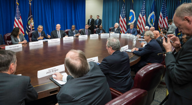 Eine Gruppe von Menschen sitzt an einem Tisch, einige stehen in der Ecke, Fahnen vor einem blauen Vorhang dahinter.