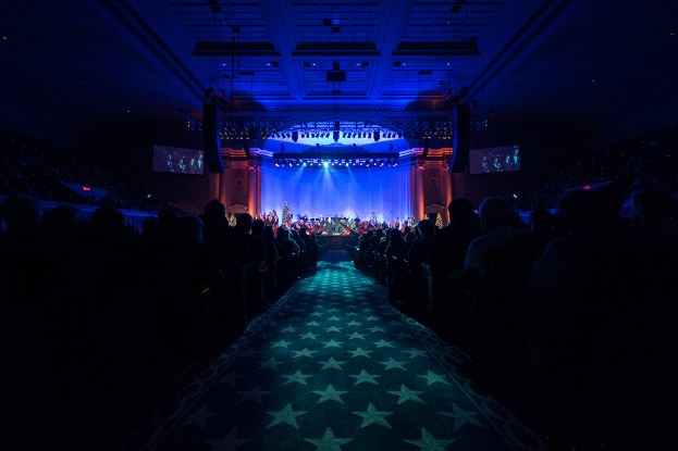 Ein großer Zuschauerraum mit Sitzenden, eine Bühne mit Musikern und eine Decke mit Lichtern und Fernsehgeräten, was auf einen Konzertsaal für die Nationale Convention 2019 hindeutet.
