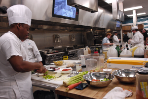 Eine Gruppe von Köchen in einer Küche beim Zubereiten von Essen, wobei ein Koch in der Mitte in einem weißen Kleid und einer Mütze steht und ein Messer hält und von verschiedenen Utensilien, Schüsseln, Gemüse, einem Handy, Flaschen und anderen Gegenständen umgeben ist, mit einem Fernseher und Deckenlampen im Hintergrund.