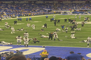 Ein Fußballspiel wird in einem Stadion gespielt, während Menschen auf dem Rasen stehen und einige auf Stühlen sitzen. Eine Person hält eine Kamera, was auf eine Live-Übertragung hinweist.