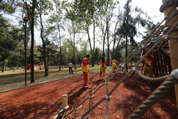 Eine Gruppe von Menschen, die einen Seilparcours in einem Park mit Bäumen im Hintergrund und einer Stange mit Seilen auf der rechten Seite absolvieren.