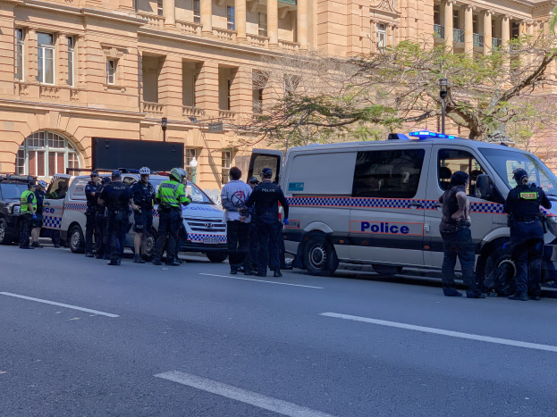 Eine Gruppe von Polizisten in Helmen steht vor einem Gebäude, mit Fahrzeugen auf der Straße und Bäumen und Laternen im Hintergrund, während einer Demonstration, die wahrscheinlich auf den Terroranschlag in Melbourne reagiert.