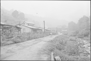 Ein Schwarz-Weiß-Foto einer Schotterstraße mit einem Pferd, das darauf läuft, umgeben von Pflanzen, Bäumen, Häusern, Pfählen, Drähten und einem Zaun, mit Hügeln und einem klaren Himmel im Hintergrund und Text am unteren Rand des Bildes.