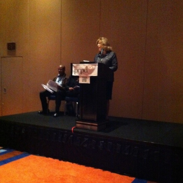 Zwei Männer sitzen auf Stühlen und eine Frau steht an einem Rednerpult auf einer Bühne, mit einer hölzernen Wand im Hintergrund und einem Teppich im Vordergrund.