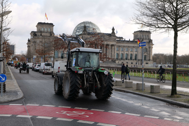 Ein grüner Traktor fährt am Reichstaggebäude in Berlin, Deutschland, vorbei, mit Fußgängern und Fahrradfahrern auf dem Gehweg, Bäumen entlang der Straße und Flaggen am Gebäude.