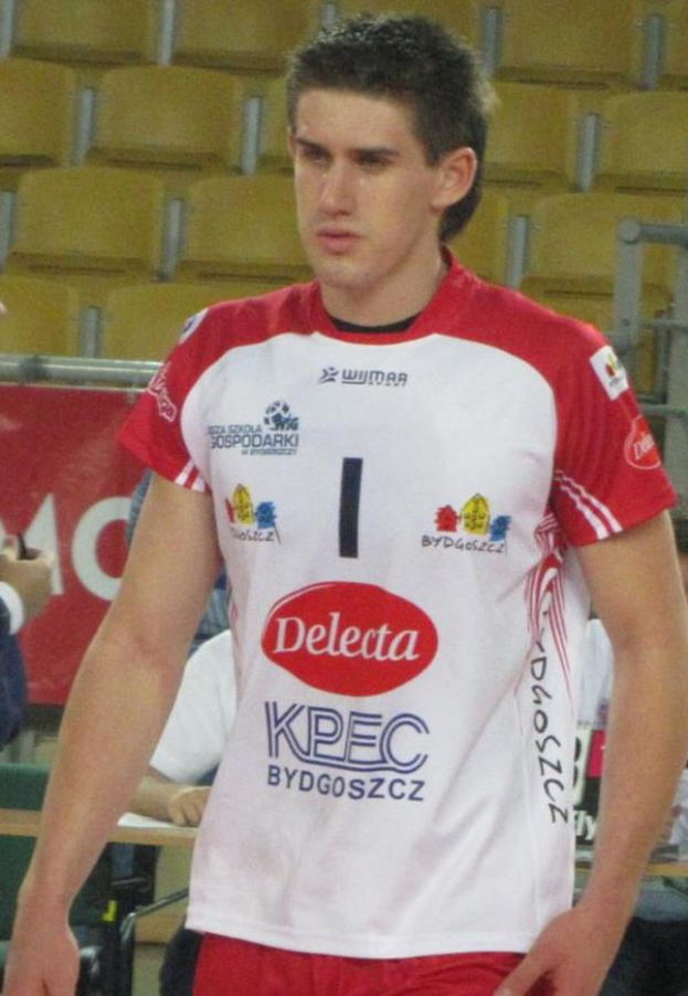 Ein Mann in rot-weißer Handballdress steht auf einem Court, umgeben von Zuschauern, mit einem roten Banner im Hintergrund, auf dem "Krasnoyarsk vs Bydgoszcz" steht, und sieht entschlossen aus.