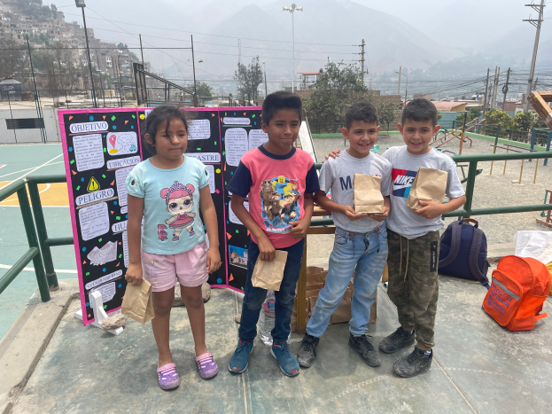 Eine Gruppe von Kindern steht auf einem Basketballfeld, jedes hält einen Papiertüten, mit Tüten und Gegenständen rechts und einer Tafel mit Text links, mit Zaun, Pfosten, Straßenlaternen, Bäumen, Gebäuden, Hügeln und Himmel im Hintergrund.