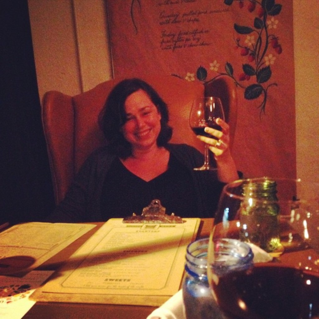 Eine Frau sitzt auf einem Stuhl, lächelt und hält ein Weinglas in der Hand, vor einem hölzernen Tisch mit verschiedenen Gegenständen, mit einem Banner, einer Pfeife und einer Wand im Hintergrund.