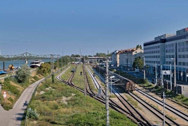 Ein Zug fährt auf Schienen neben einer Stadtlandschaft mit Strommasten, Gebäuden, Grünflächen, Straßenschildern, Fahrzeugen, einer Brücke, Wasser und einem bewölkten Himmel.