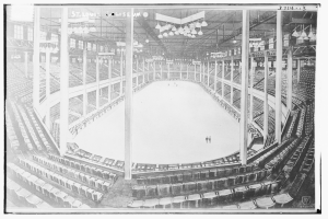 Ein altes Schwarz-Weiß-Foto einer Eishalle mit Menschen, die Schlittschuh laufen, Stühlen, Säulen, Lampen und einer Decke, mit Text unten.