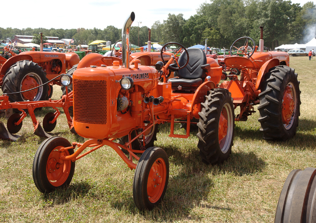 Mehrere orangen Traktoren auf einem Rasen mit Gras im Vordergrund, verschiedene Fahrzeuge, grüne, blaue und weiße Schirmzelte, Bäume und Gebäude im Hintergrund und zwei Männer auf der rechten Seite unter einem klaren Himmel.
