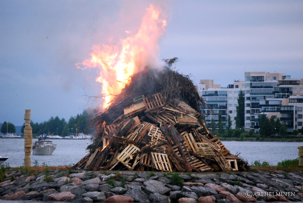Holzobjekte brennen mit Feuer, Steine unten, ein Schiff auf dem Wasser in der Mitte, mit Gebäuden, Bäumen und Himmel im Hintergrund.