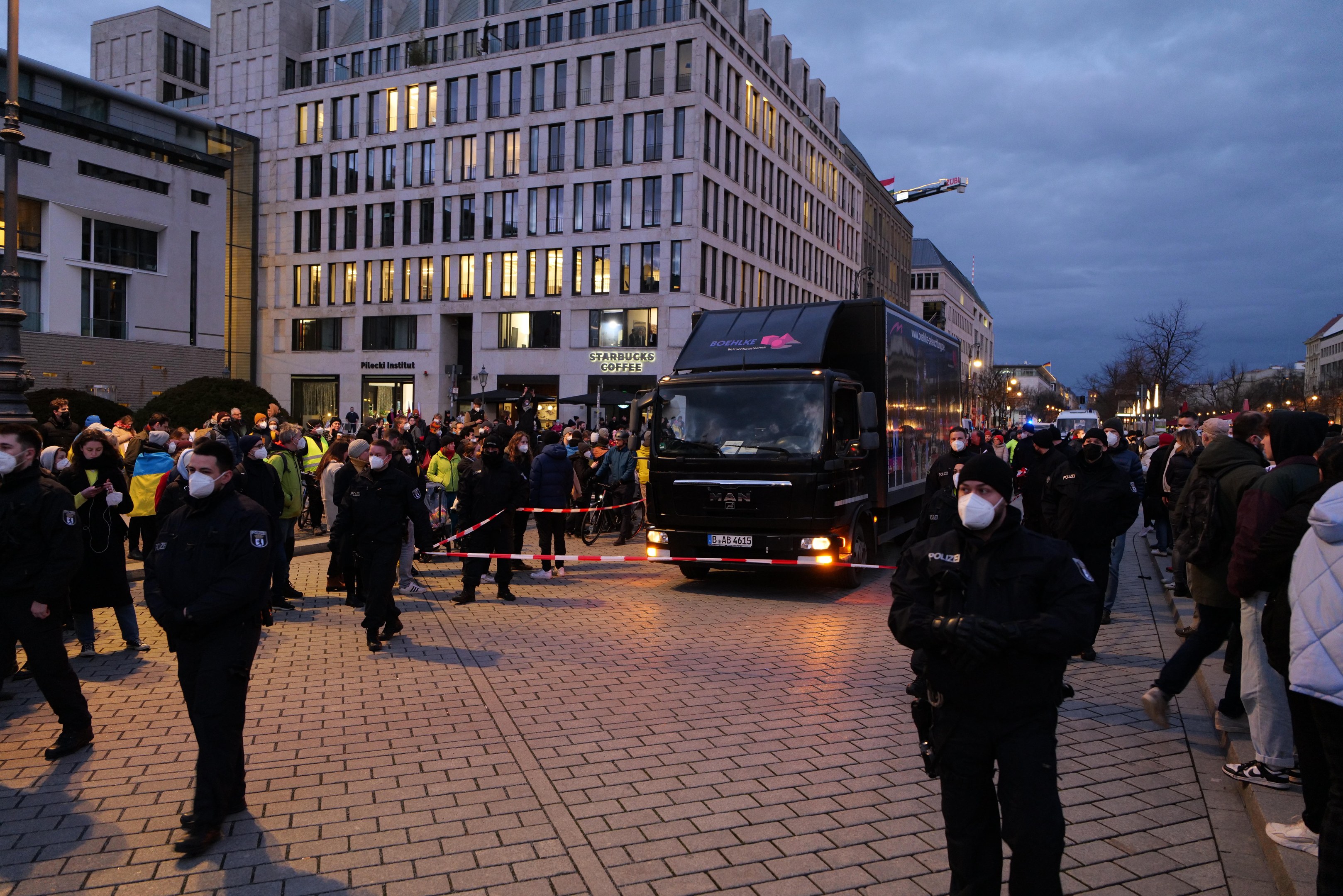 Eine Gruppe von Menschen steht in der Nähe eines Lastwagens auf einer Straße, die von Gebäuden, Bäumen und Laternen gesäumt ist, unter einem bewölkten Himmel, wobei einige Mützen und Masken tragen und ein Band auf einem Pfahl im Vordergrund.