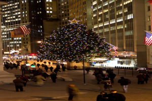 Ein Weihnachtsbaum mit Menschen darauf in der Mitte einer Straße, mit zwei Fahnenmasten auf beiden Seiten und beleuchteten Gebäuden im Hintergrund.