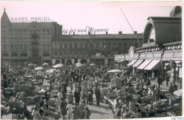 Schwarzes und weißes Foto eines belebten Berliner Markts mit Menschen, Gemüsewagen, Schirmen und Gebäuden im Hintergrund.