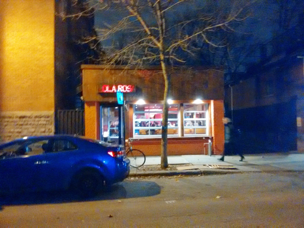 Eine nächtliche Szene mit einem Auto auf einer Straße, einer Person und einem Fahrrad auf einem Fußweg neben der Straße, einem Baum, einem Geschäft und einigen Gebäuden in der Mitte.