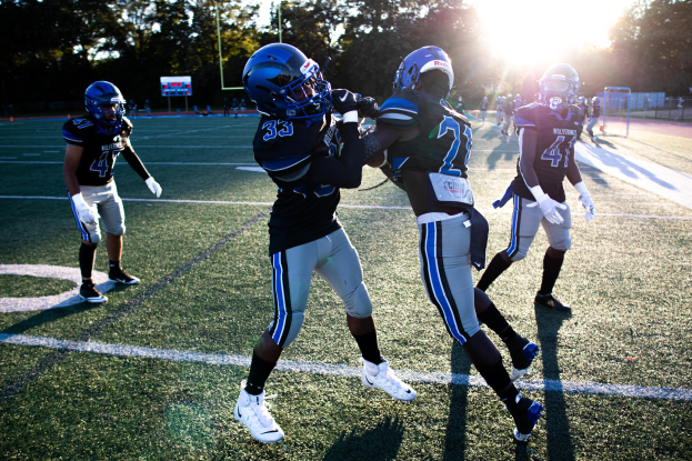 Gruppe junger Männer beim Fußballspielen auf einem Feld mit Bäumen und einem klaren blauen Himmel, die Helme und Schuhe tragen.