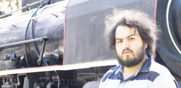 Ein Mann in einem T-Shirt steht neben einem Zug, wobei das Zugrad und andere Teile unten sowie ein Baum in der oberen linken Ecke zu sehen sind.
