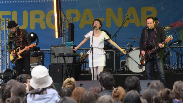 Vier Personen auf der Bühne: Eine Frau in einem weißen Kleid singt in der Mitte, flankiert von zwei Männern, die Gitarren spielen, während eine Menge darunter eine Wand mit Lautsprechern im Hintergrund betrachtet.