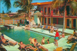 Ein Schwimmbad mit Menschen auf Stühlen im Vordergrund, mit Gebäuden, Bäumen und Autos im Hintergrund.