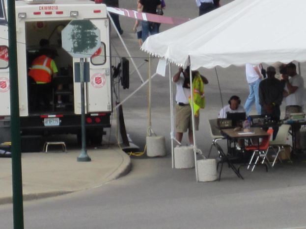 Ein Rettungswagen befindet sich auf der linken Seite einer Stra├če, w├Ąhrend auf der rechten Seite ein Zelt eine Gruppe von Menschen sch├╝tzt, mit einem Tisch und einem Stuhl in der N├Ąhe.