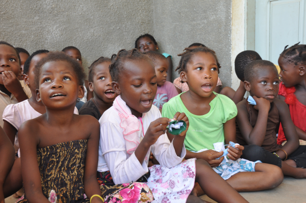 Eine Gruppe von Kindern sitzt auf dem Boden vor einem Gebäude, mit einer Wand und einer Tür im Hintergrund, mit traurigen und verzweifelten Gesichtern.