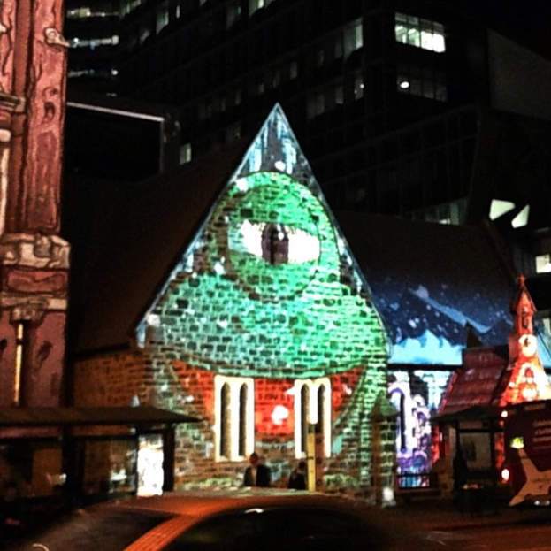 Ein zentrales Haus mit einer Person in der Nähe, flankiert von Häusern auf beiden Seiten, ein Gebäude und Lichter im Hintergrund und ein Auto unten.