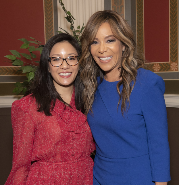 Zwei Frauen in Kleidern stehen lachend nebeneinander vor einer Vorhangwand, mit einer Pflanze im Hintergrund.