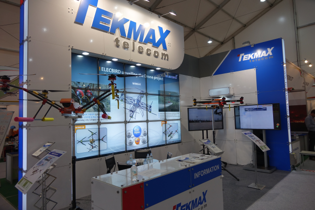Ein Messestand von Tekmax Telecom mit einer großen Wandanzeige, einem Tisch mit Produkten, zwei Fernsehern, Informationsplakaten, Deckenleuchten und einigen Personen.