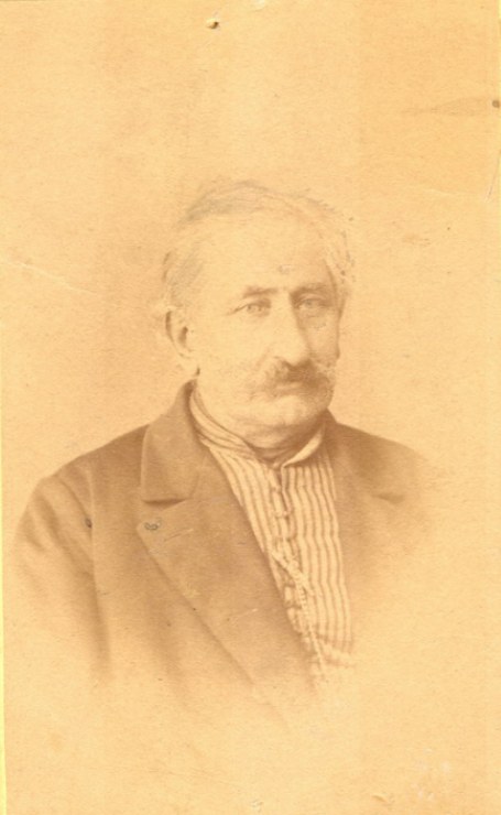 Ein Schwarz-Weiß-Porträt eines älteren Mannes mit Schnurrbart, der einen Mantel trägt, mit markantem Kinn, großer Nase, dunklen Augen und einem nachdenklichen Ausdruck mit leicht geöffneten Lippen.