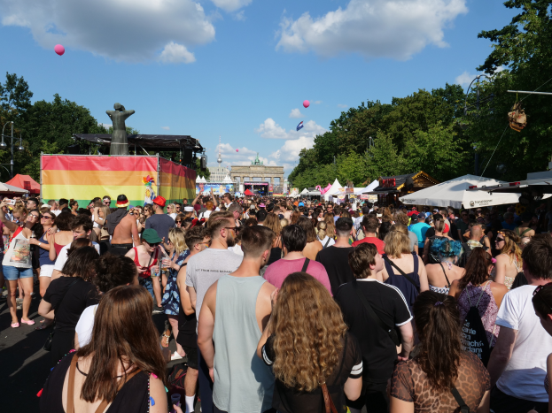 Eine große Menschenmenge, die eine Straße mit Zelten, Bäumen, Pfählen und Lichtern entlanggeht, mit Gebäuden und einem bewölkten Himmel mit Ballons im Hintergrund beim Christopher Street Day in Berlin.