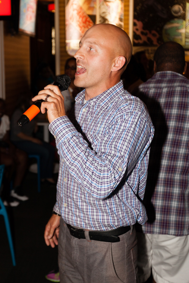 Eine Person in einem schwarzen Gürtel steht und singt in ein Mikrofon mit einer Gruppe von Menschen hinter sich.