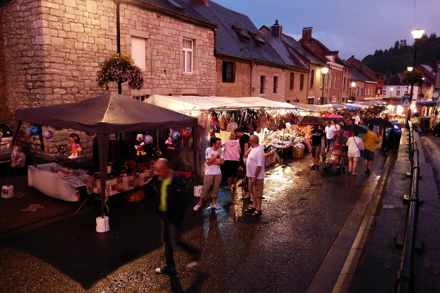 Eine Gruppe von Menschen, die auf einer Straße mit Geschäften, Schirmen, hängenden Spielzeugen, Taschen, Gebäuden mit Fenstern, Bäumen, Straßenlaternen und Barrikaden geht.