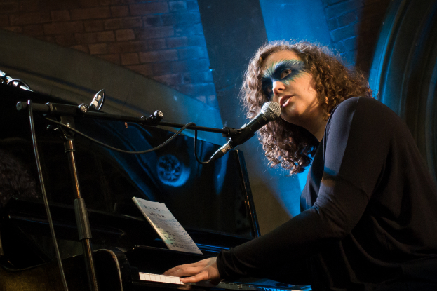 Eine Frau mit kurzen braunen Haaren sitzt an einem Piano und schaut zur Seite, während sie in ein Mikrofon singt.