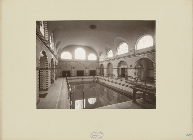 Ein Schwarz-Weiß-Foto eines Schwimmbads im Innenraum, umgeben von Geländern, Bänken, Bögen und Wandlampen, mit Text unten.