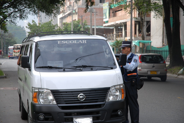 Ein Polizeibeamter spricht mit einer Person in einem Fahrzeug auf einer vielbefahrenen Straße umgeben von Bäumen, hellbraunen Gebäuden und Strommasten.