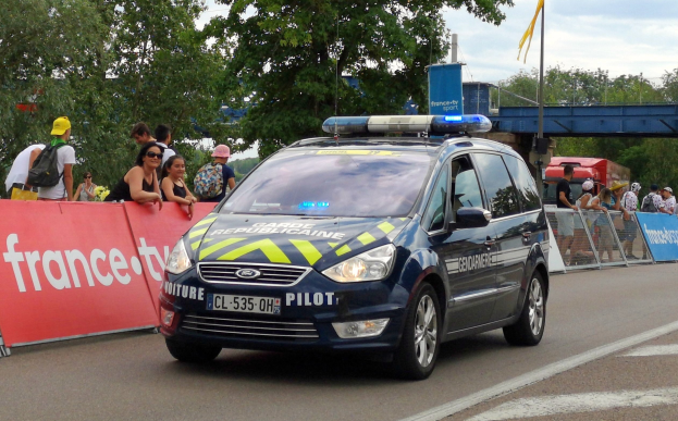 Polizeiauto fährt an einer Menschenmenge mit Schildern, Geländern, Bäumen, einer Brücke, einer Flagge und einem bewölkten Himmel vorbei.