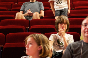 Eine Gruppe von Menschen in einem Kino mit einem Jungen in der Mitte, der ein weißes T-Shirt und schwarze Hosen trägt.